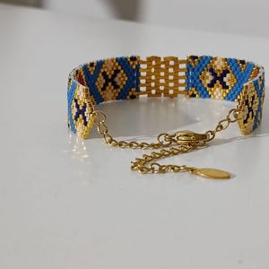 May include: A beaded bracelet with a gold chain and clasp. The bracelet features a geometric pattern in blue, tan, and dark blue beads. The bracelet is approximately 1 inch wide.