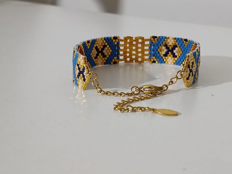 May include: A beaded bracelet with a gold chain and clasp. The bracelet features a geometric pattern in blue, tan, and dark blue beads. The bracelet is approximately 1 inch wide.