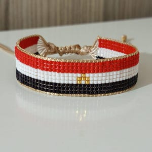 Puede incluir: Una pulsera de cuentas con los colores de la bandera de Egipto. La pulsera es roja, blanca y negra con un emblema dorado en el centro.