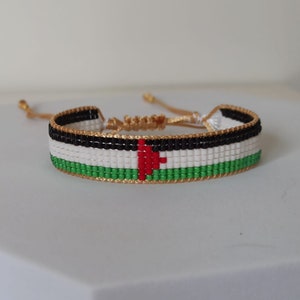 May include: A beaded bracelet with the colors of the Palestinian flag. The bracelet is made with black, white, red, and green beads. The bracelet has a gold cord.