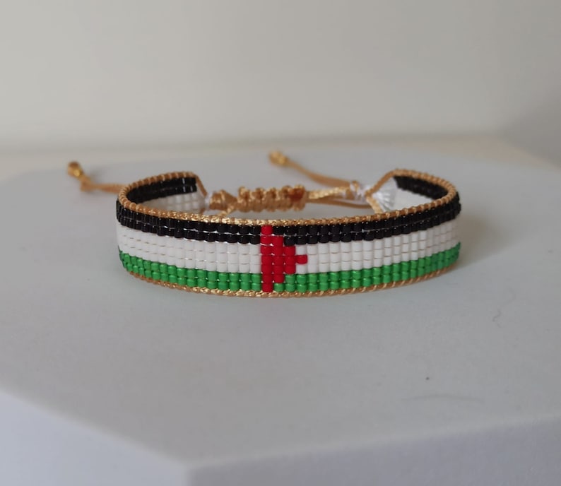 May include: A beaded bracelet with the colors of the Palestinian flag. The bracelet is made with black, white, red, and green beads. The bracelet has a gold cord.