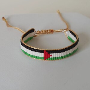 May include: A woven bracelet with the colors of the Palestinian flag. The bracelet is made with green, white, black, and red thread. It has a gold clasp.