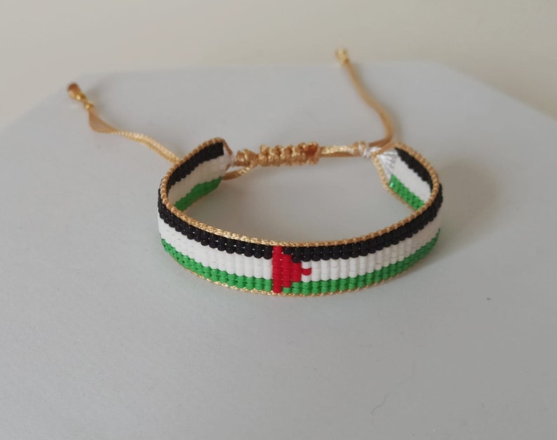 May include: A woven bracelet with the colors of the Palestinian flag. The bracelet is made with green, white, black, and red thread. It has a gold clasp.