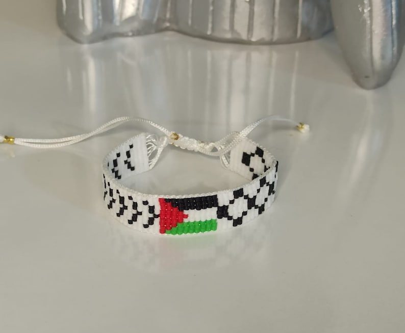 May include: White beaded bracelet with a black and white checkered pattern and a small Palestinian flag in the center.