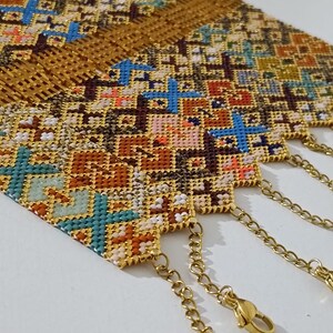 May include: Close-up of several beaded bracelets with intricate cross-stitch patterns in shades of gold, brown, blue, and orange. Each bracelet has a gold chain and clasp for closure. The bracelets are displayed on a white surface.