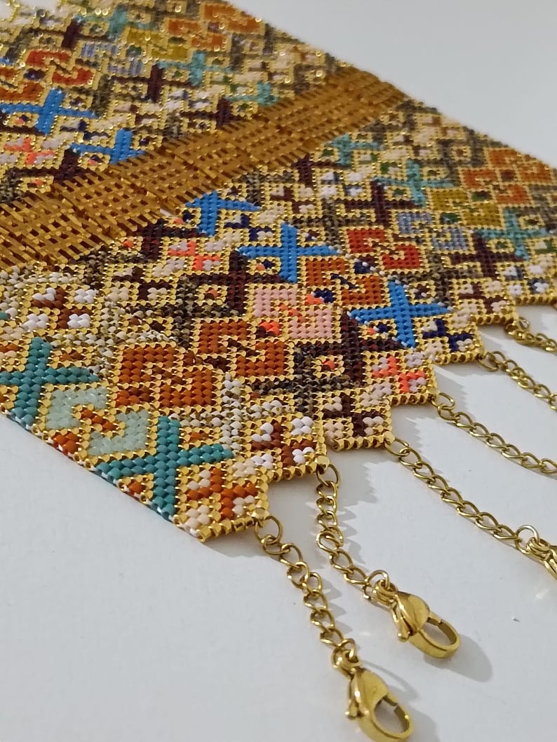 May include: Close-up of several beaded bracelets with intricate cross-stitch patterns in shades of gold, brown, blue, and orange. Each bracelet has a gold chain and clasp for closure. The bracelets are displayed on a white surface.