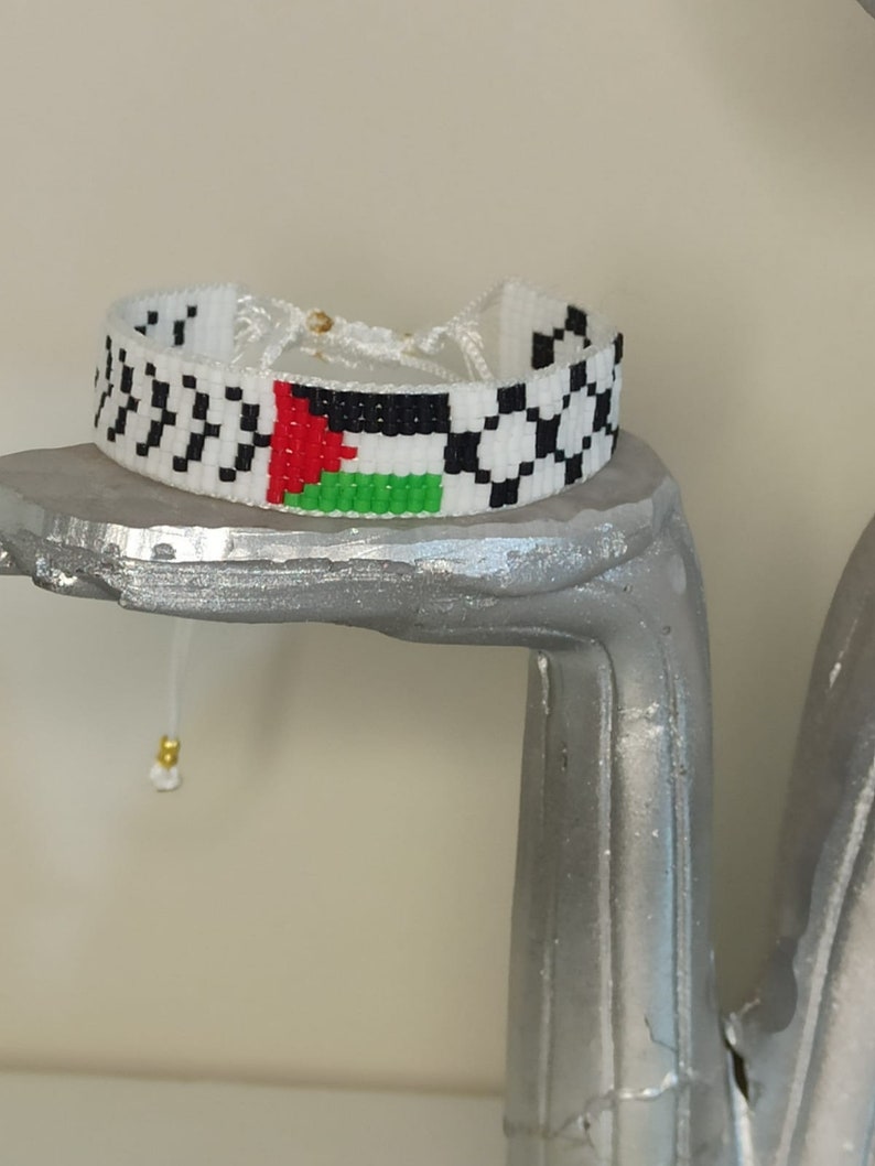 May include: A white beaded bracelet with a black and white checkered pattern on either side of a Palestinian flag.