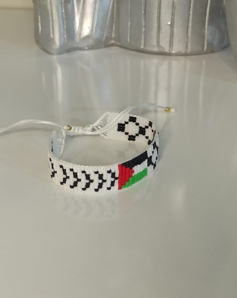 May include: White beaded bracelet with a black and white pattern and a small Palestinian flag design.