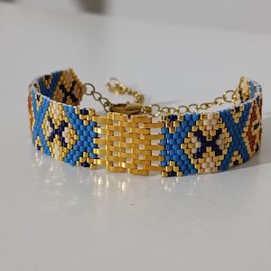 May include: A beaded bracelet with a gold-colored clasp and chain. The bracelet features a geometric pattern in blue, gold, cream, and brown beads. The central portion of the bracelet is a gold-colored woven design.