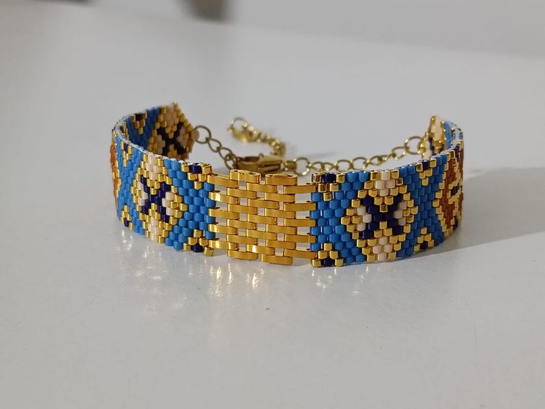 May include: A beaded bracelet with a gold-colored clasp and chain. The bracelet features a geometric pattern in blue, gold, cream, and brown beads. The central portion of the bracelet is a gold-colored woven design.