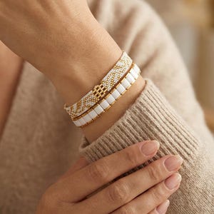 Op de afbeelding: Twee kralenarmbanden zijn op een pols gestapeld. De bovenste armband heeft een goud-wit geometrisch patroon. De onderste armband is wit met gouden accenten. De armbanden zijn gemaakt van kleine kralen.