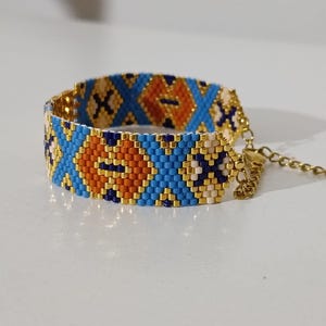 May include: A beaded bracelet with a geometric pattern in blue, orange, and white. The bracelet has a gold-colored chain and clasp. The design features diamond and X shapes.