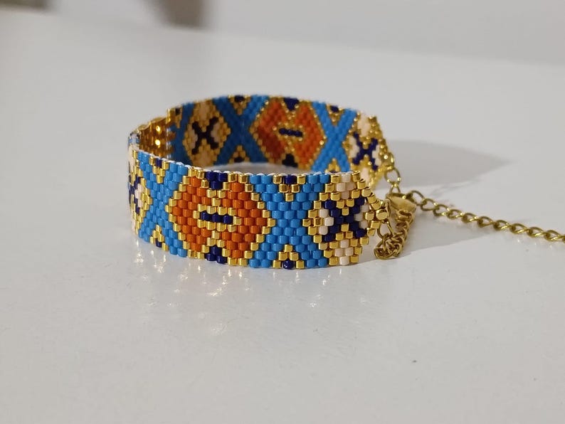 May include: A beaded bracelet with a geometric pattern in blue, orange, and white. The bracelet has a gold-colored chain and clasp. The design features diamond and X shapes.