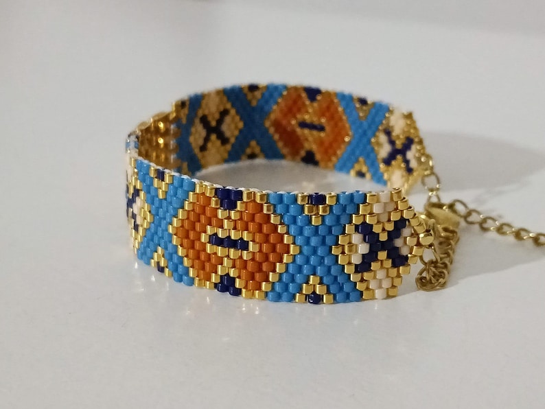 May include: A beaded bracelet with a geometric pattern. The bracelet features a repeating design of blue, orange, and gold beads. The bracelet is adjustable and has a gold-colored chain.