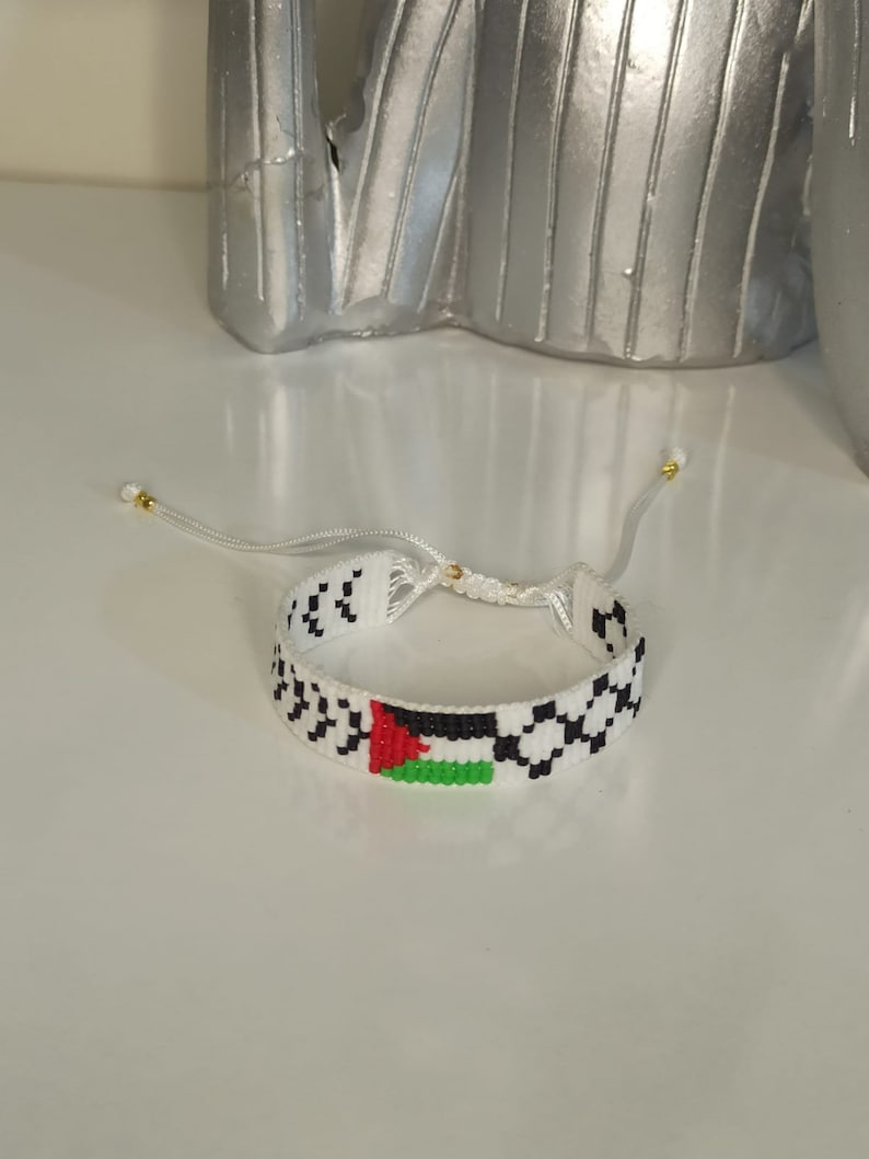 May include: White beaded bracelet with a woven pattern and a small Palestinian flag in red, black, green, and white.
