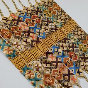 May include: Close-up of several gold chain necklaces with intricately woven, colorful patterns. The patterns feature geometric shapes in shades of brown, blue, orange, and white. The necklaces are displayed on a white surface.