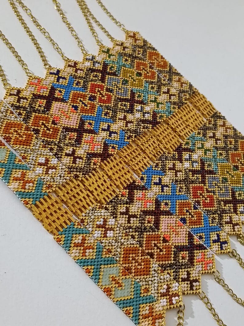 May include: Close-up of several gold chain necklaces with intricately woven, colorful patterns. The patterns feature geometric shapes in shades of brown, blue, orange, and white. The necklaces are displayed on a white surface.