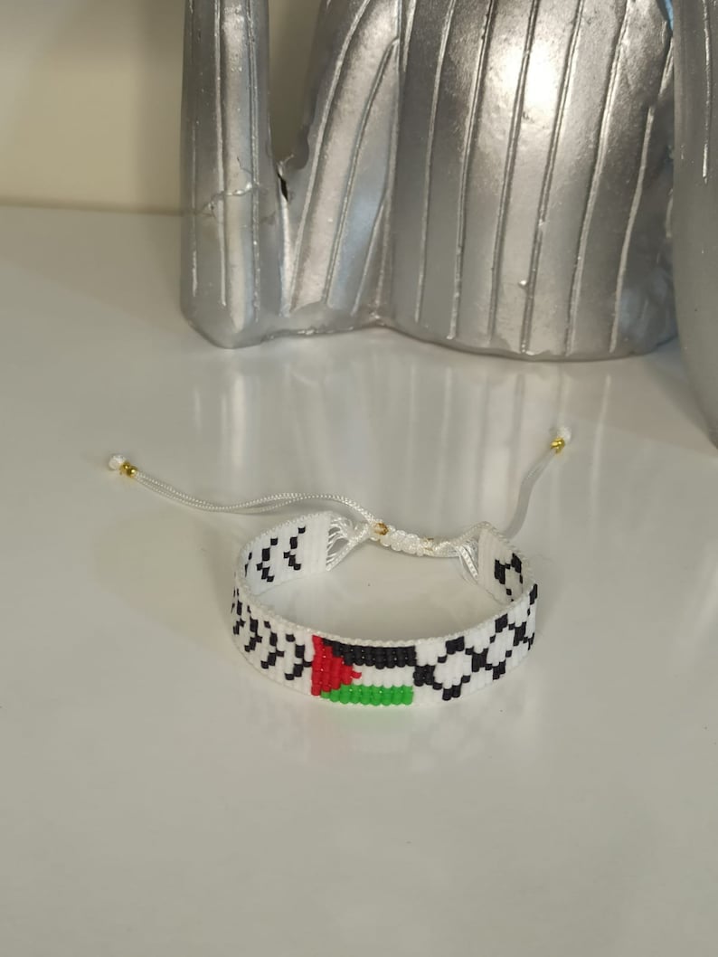 May include: White beaded bracelet with a black and white checkered pattern and a small Palestinian flag in the center.