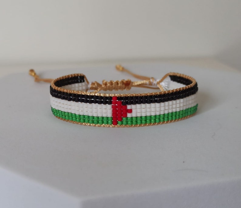 May include: A beaded bracelet with the colors of the Palestinian flag. The bracelet is made with small, colorful beads and has a gold cord closure.