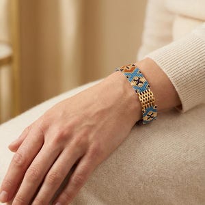 May include: A beaded bracelet with a gold-tone chain and a colorful pattern of blue, orange, and black beads. The bracelet is worn on a wrist, showcasing the intricate design and craftsmanship. The bracelet's design features geometric shapes.