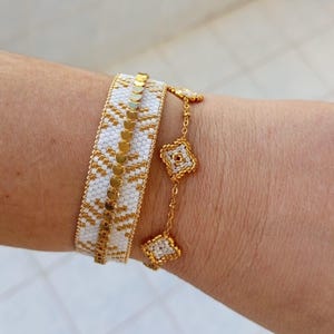 以下が含まれることがあります： 金と白のビーズブレスレット2本。1つは金のアクセントが付いた幅広のカフで、もう1つは正方形とひし形の金と白のビーズが付いた繊細なチェーンブレスレットです。ブレスレットは手首に着用されています。