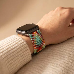 May include: A smartwatch with a black rectangular screen and a woven watch strap featuring a multi-coloured geometric pattern in shades of turquoise, red, yellow, and white. The strap is constructed from small beads or threads in a chevron design.