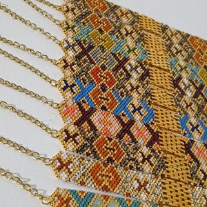 May include: Close-up of several beaded bracelets with gold-colored chain clasps. The bracelets feature intricate geometric patterns in various colors, including orange, blue, white, and brown. The beads are tightly woven together, creating a textured surface.