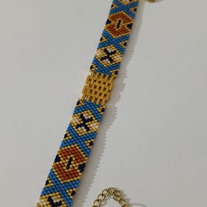 May include: A beaded bracelet with a gold clasp and extender chain. The bracelet features a pattern of blue, gold, white, and brown beads. The clasp and chain are gold-colored. The bracelet is approximately 7 inches long.