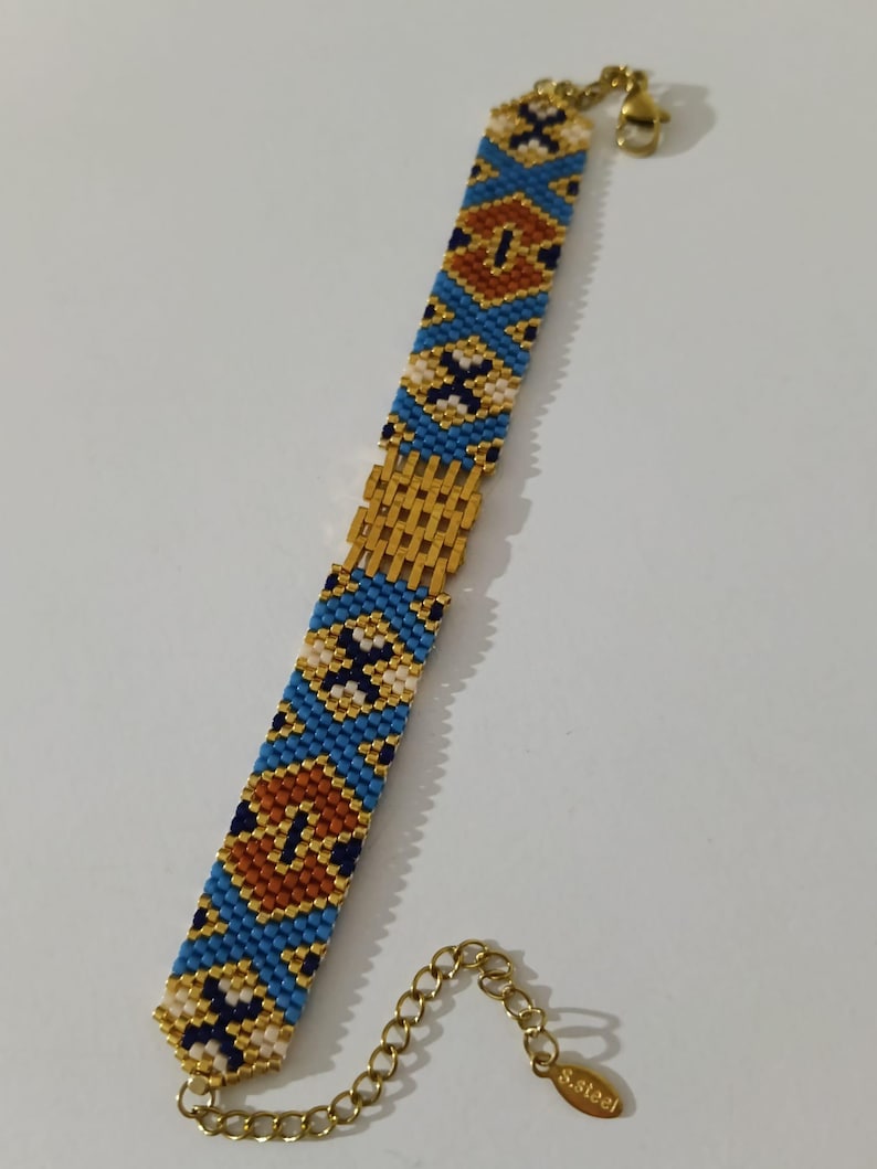 May include: A beaded bracelet with a gold clasp and extender chain. The bracelet features a pattern of blue, gold, white, and brown beads. The clasp and chain are gold-colored. The bracelet is approximately 7 inches long.