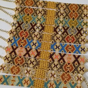 May include: A collection of beaded bracelets with intricate geometric patterns in shades of brown, gold, blue, and orange. Each bracelet features a gold chain clasp. The bracelets are displayed side by side.