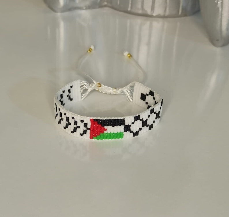 May include: White beaded bracelet with a black and white checkered pattern and a Palestinian flag in the center.