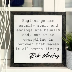 May include: Square wooden sign with a dark gray frame. The white background features black text that reads "Beginnings are usually scary and endings are usually sad, but it is everything in between that makes it all worth living." Signed "-Bob Marley."