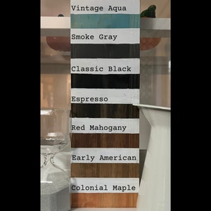 Może przedstawiać: Drewniana paleta kolor&oacute;w prezentująca osiem kolor&oacute;w farb: Vintage Aqua, Smoke Gray, Classic Black, Espresso, Red Mahogany, Early American i Colonial Maple. Każdy kolor jest oznaczony białym tekstem na białym tle. Biały dzbanek i klepsydra znajdują się w tle.