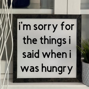 May include: Square wooden sign with a white background and black text that reads "I'm sorry for the things I said when I was hungry." The sign has a dark gray frame and is displayed on a white surface.