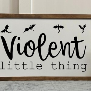 May include: Wooden framed sign with the words "Violent little thing" in black script. Above the text are four black dragon silhouettes. The sign is on a white background with a brown frame. Decorative home decor.