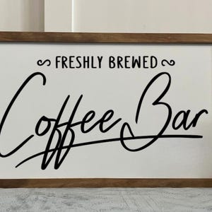 May include: A rectangular wooden framed sign with the words "FRESHLY BREWED Coffee Bar" in black script. The sign is on a white background. A decorative white pitcher and a small plant are in the background.