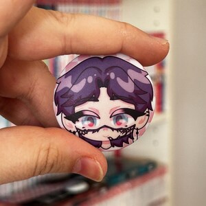 Op de afbeelding: Een ronde button met een anime-stijl karakter met paars haar en opvallende blauwe ogen. Het karakter heeft gedetailleerde zwarte accenten rond de ogen en mond, tegen een roze en witte achtergrond.