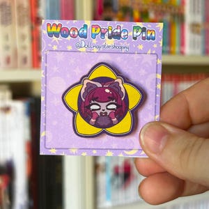 Könnte beinhalten: Ein Wood Pride Pin mit einem Cartoon-Charakter-Design, auf einer lila Rückkarte. Der Pin hat eine Blumenform mit gelbem Umriss. Auf der Karte steht "Wood Pride Pin" und der Benutzername des Verkäufers. Der Pin wird von einer Hand gehalten.