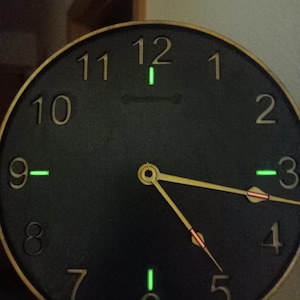 May include: A black clock face with gold numbers and hands. The clock has a wooden frame and glows green in the dark.