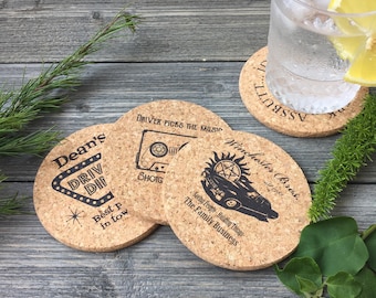 May include: Three round cork coasters with black text and images. The first coaster says "Dean's" with a neon sign that says "Drive Thru". The second coaster says "Driver Picks the Music" with a cassette tape and the word "Shotg". The third coaster says "Winchester Bros. est. 1983" with a car and a pentagram. The text below the car says "Saving People Hunting Things The Family Business".