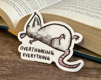May include: A white sticker with a black and gray cartoon rat lying on its back with its legs in the air. The text "Overthinking Everything" is written below the rat.