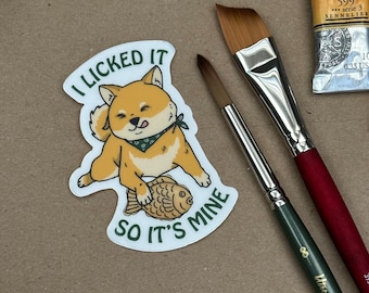 May include: A sticker featuring a cartoon Shiba Inu dog wearing a bandana, holding a fish-shaped taiyaki pastry. The sticker reads "I licked it so it's mine".