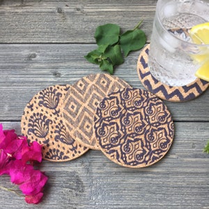 May include: Four round cork coasters with different geometric and floral patterns in black and white on a wooden surface.