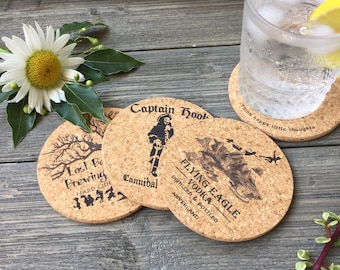 May include: Four round cork coasters with different designs. The coasters feature black and white illustrations of a pirate, a tree, and a ship. The text on the coasters reads "Lost Boy Brewing Hard Cider", "Captain Hook Cannibal", "Flying Eagle Vodka Distilled & Bottled Neverland", and "Think happy little thoughts".