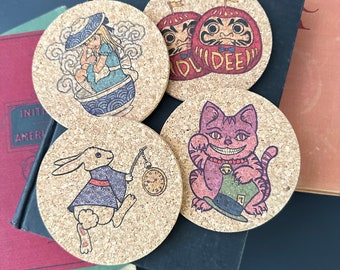 May include: Four cork coasters with different designs. One features a girl in a blue dress and a bowl with a wave pattern. Another has a white rabbit with a pocket watch. The third has a red cat with a green hat and the text "Mad Dee". The last one has two red daruma dolls with the text "Mad Dee".