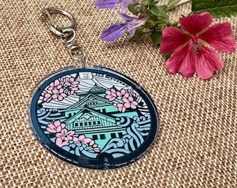 May include: A round, clear acrylic keychain with a black and white image of a Japanese castle with pink cherry blossoms. The keychain is attached to a silver metal ring.