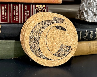 May include: Cork coaster with a black and white doodle design of the letter C. The letter is surrounded by a variety of small doodles, including animals, plants, and geometric shapes.