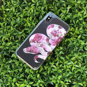 May include: A black phone case with a floral design and a snake wrapped around the flowers. The snake is pink and white with black eyes. The flowers are pink and white.