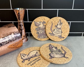 May include: Four cork coasters with humorous rat illustrations and text. One rat is in a trash can with the text "EATING MY EMOTIONS". Another says "SINCERE APOLOGIES FOR MY SOCIAL AWKWARDNESS". The third says "OVERTHINKING EVERYTHING". The fourth says "ANXIETY IS MY CHILDO".