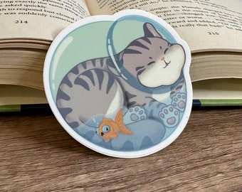 May include: A die-cut sticker featuring a cartoon cat inside a bubble, curled up and sleeping. The cat is gray with darker stripes, and a small orange fish swims below. The bubble is light blue, and the sticker is on a book.